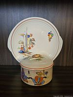 Covered jar with lid in front of matching serving plate displaying folk scene design