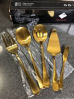 Five gold-tone stainless steel serving utensils displayed beside original box