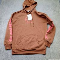 Full view of brown hoodie front, showing graphic, sleeve text, pocket, hood, and attached tag