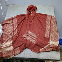 Full view of rust and beige striped hooded poncho laid flat showing hood, stripes and fringed hem