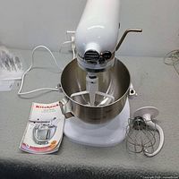 KitchenAid stand mixer with bowl, flat beater installed, wire whisk attachment and manual visible