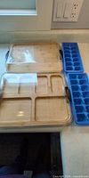 Two bamboo sectioned serving trays with clear lids alongside two blue silicone ice cube trays