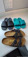 Three pairs of sandals laid out: black Birkenstock strappy pair, black Crocs slides, teal Crocs slides