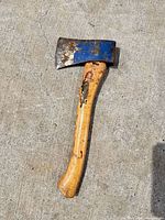 Full view of hatchet lying on concrete, showing wooden handle, blue-painted steel head and overall wear