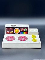 Front view showing full cooktop with burners, dial, knobs, and Fisher-Price panel