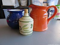 Front view of three ceramic items: orange pitcher, blue pitcher, olive oil bottle