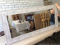 Mirror laid horizontally on a padded surface, showing white grey distressed wooden frame, reflective glass, and surrounding background including table and chairs.
