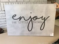 Photo shows a 24x36 inch rectangular wall art piece with a grey textured background and black script reading 'Enjoy The Little Things'. Held upright inside a room near a fireplace.