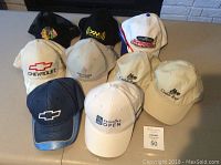 Photo showing front view of 9 caps arranged in rows featuring different logos and colors including Chevrolet, Cedar Run, Canadian Open, and Power Race logos clearly visible.