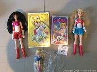 Photo showing two vintage Sailor Moon dolls, a smaller doll still sealed, and two VHS tapes with Sailor Moon themes.