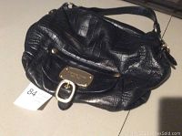 Front view of Michael Kors black leather purse showing textured leather, gold tone front metal plate and buckle, and strap with slightly worn edges.