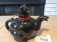 Front angled view of black ceramic cat teapot with red collar and green eyes, spout shaped like paw, handle as tail.