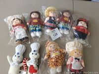 Seven Campbell's Soup soft dolls and two Pillsbury Christmas Doughboy soft dolls arranged on a flat surface, all in original plastic bags.