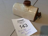 Side view of vintage ceramic miniature hot water bottle warmer showing light cream body with brown accents and screw stopper.