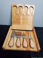 Open wooden case displaying four wood-handled cheese tools seated in carved recesses
