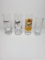 Four tall shooter glasses lined up: Cazadores, Sauza black logo, Sauza Gold, unbranded tall glass
