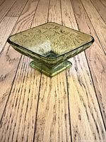 Front view of square green pressed glass pedestal bowl on wood floor