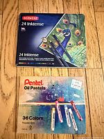 Derwent Inktense tin above Pentel oil pastel box on wooden floor