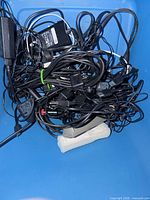 Tangled grouping of black cables, several plug-in adapters, and a white six-outlet power bar in a blue bin