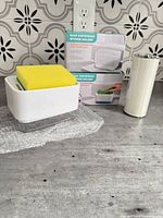 Countertop view of unboxed soap dispensing sponge holder with sponge, two boxed units, and rolled silicone faucet mat