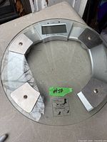 Top view of round glass digital scale showing sensor pads and battery warning label