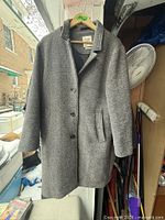 Front view of grey herringbone coat hanging, showing three-button closure and pockets