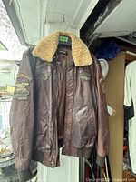 Full front view of brown leather bomber jacket hanging, showing shearling collar and patches