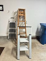 Overall view showing 2-step stool in front, wood ladders stacked, 3-step stool at back