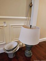 Lamp with drum shade; two additional shades stacked beside lamp; curtain rod leaning against wall