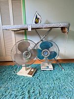 Ironing board standing upright with two oscillating fans beneath