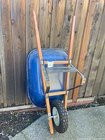 Wheelbarrow leaned against fence, side view showing blue tray, wooden handles, tire