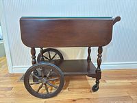 Side view of tea cart showing one drop leaf down, large spoked wheel, caster wheel, lower shelf, dark brown finish