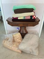 Stacked crocheted afghans on table with two bagged comforters on floor