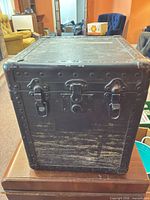 Front view of black steamer trunk showing riveted edges, latches, lock, and surface wear