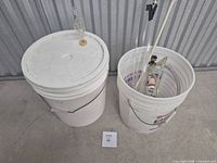Two white plastic buckets, fermenter on left with airlock, bottling bucket on right containing tubing and tools