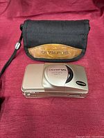 Front view of Olympus Stylus Zoom 115 camera beside Olympus case