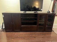 Front view of dark brown wooden media console with TV on top; shows overall size, left wood doors, central open shelves and drawer, right glass doors