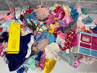 Pile of assorted Barbie doll clothing with yellow locker and pink-blue furniture piece visible
