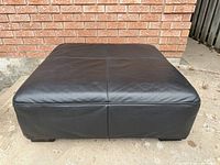 Front angled view of black leather ottoman showing stitched top and feet