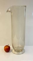 Full view of tall clear cylindrical glass pitcher beside apple for scale