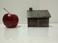Front view of house-shaped coin bank next to apple for scale, shows repoussé figures and overall condition