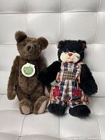 Front view of both plush toys: brown bear with tag, black cat in plaid overalls