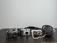 Group shot of three digital cameras, neck strap, and Optex case
