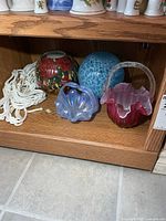 All five items on shelf: macrame hanger, red-green vase bowl, iridescent blue dish, blue crackle solar ball, pink-white basket