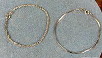 Both bracelets laid flat on fabric showing chain styles and clasps