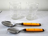 two clear glass tumblers and two yellow-handled stainless steel spoons