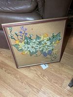 Full view of framed floral needlepoint artwork leaning against furniture