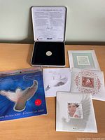 Open presentation box showing coin, certificate text in lid, and three stamp sheets beside box along with outer folder