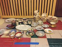 Full assortment of assorted china laid out on floor, featuring multiple teacups, saucers, plates, and serving dishes with various floral designs and colors including gold detailing.