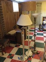 Three floor lamps standing on a multicolored tiled floor near wood paneled walls, two lamps have fabric shades and one has plastic torchiere shade.
